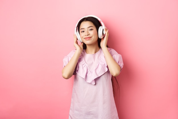 Belle femme asiatique écoutant de la musique sur des écouteurs sans fil, souriant heureux à la caméra, debout en robe sur fond rose.