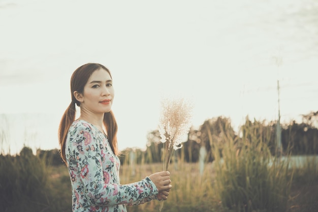 Belle femme asiatique garder la fleur d'herbe dans les champs avec un soleil éclatant, elle très heureusestyle vintagetons sombrescoucher du soleil