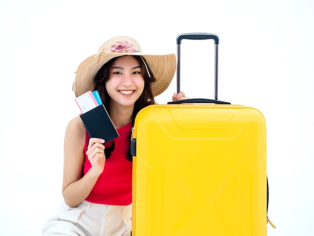 Belle femme asiatique heureuse en chemise sans manches rose et chapeau de plage sourire et tenant passeport billet d'avion et valise jaune isolé sur fond blanc Jeune voyageur ou touriste prêt à voyager