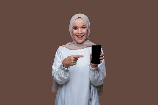 Belle femme asiatique en hijab montre un téléphone portable et pointe vers l'écran du téléphone avec un sourire