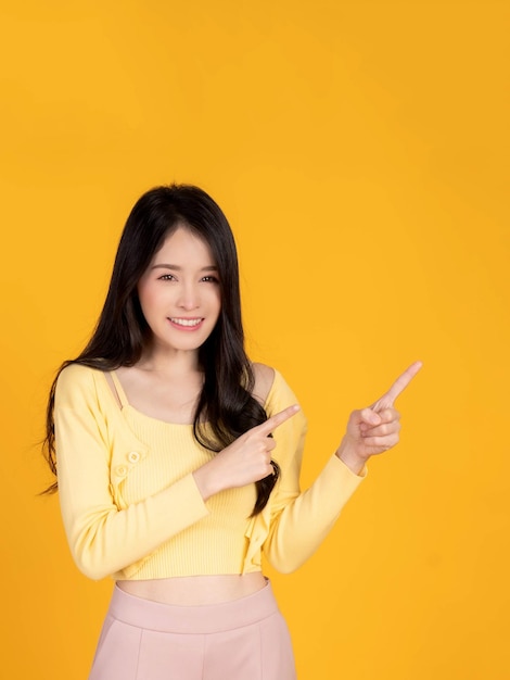 Belle femme asiatique jolie fille avec une chemise jaune souriante et montrant du doigt à l'espace de copie vide pour le produit présent ou l'espace vide pour la publicité isolé sur fond jaune
