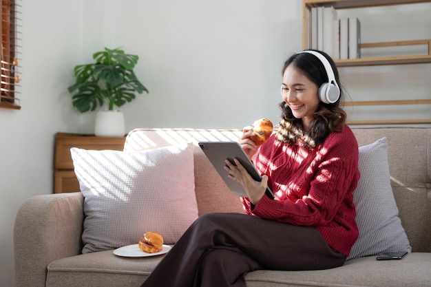 Belle femme asiatique portant un chandail écoutant de la musique en ligne avec une tablette assise sur un canapé dans le