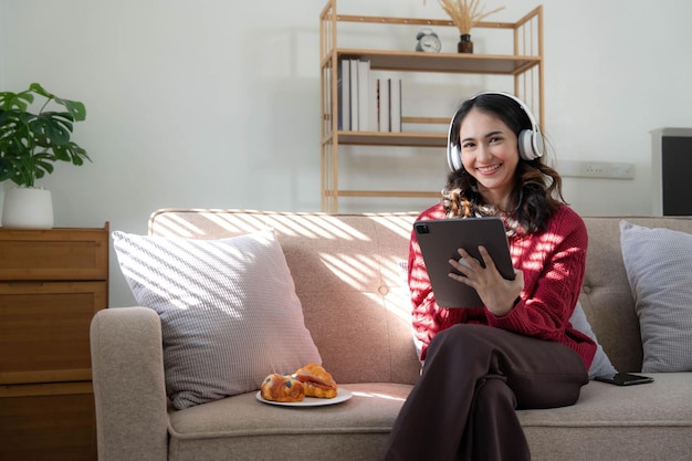 Belle femme asiatique portant un chandail écoutant de la musique en ligne avec une tablette assise sur un canapé