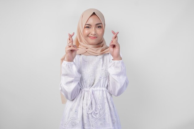 Belle femme asiatique portant un hijab et une robe blanche avec des doigts posant un souhait de chance geste debout sur un fond blanc isolé concept de Ramadan et Eid Mubarak