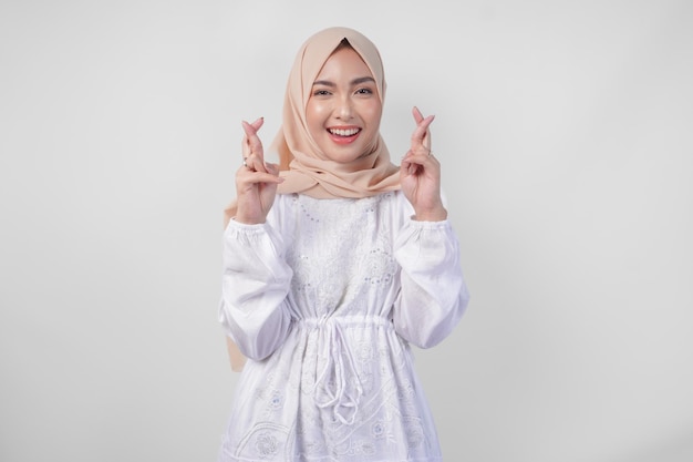 Belle femme asiatique portant un hijab et une robe blanche avec des doigts posant un souhait de chance geste debout sur un fond blanc isolé concept de Ramadan et Eid Mubarak