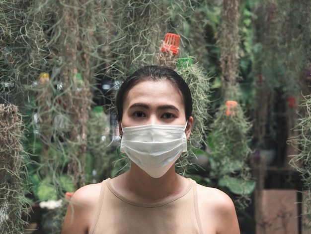 Photo belle femme asiatique portant un masque médical propre et blanc et une chemise sans manches beige et regardant la caméra dans le jardin verdoyant. heureux et amoureux de la nature, nouveau concept normal.