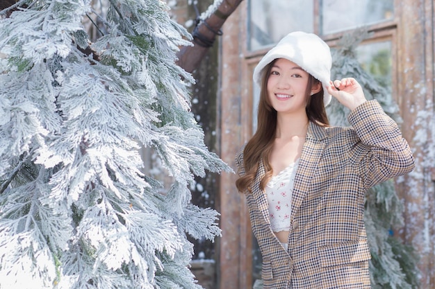 Belle femme asiatique porte un chapeau blanc et un manteau à carreaux et se tient près de la neige recouverte d'un arbre de Noël