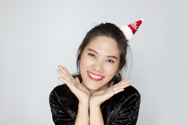 Une belle femme asiatique porte un petit chapeau de Noël rouge joyeux et lève la main de la paume sous le menton avec le sourire et le bonheur pour célébrer Noël et bonne année