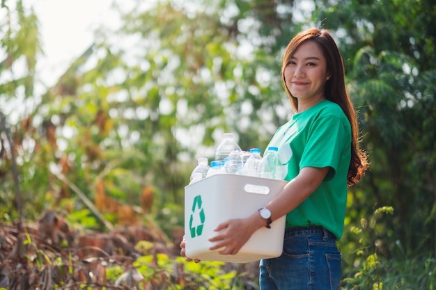 Une belle femme asiatique ramassant des ordures et tenant une corbeille avec des bouteilles en plastique à l'extérieur