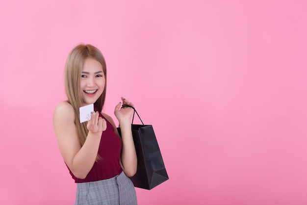 Belle femme asiatique avec sac à provisions et carte de crédit à la main sur fond roseLes Thaïlandais utilisent une carte de crédit et aiment faire du shopping