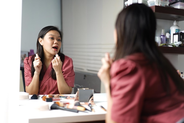 Belle femme asiatique se maquiller et appliquer du rouge à lèvres devant un miroir de coiffeuse