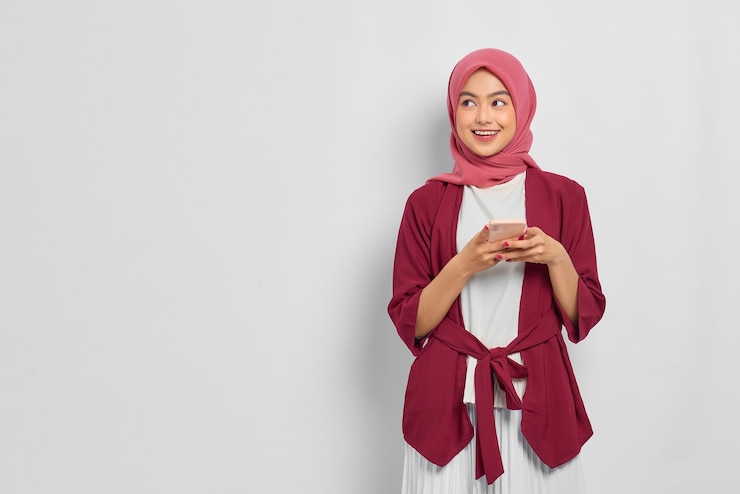 Belle femme asiatique souriante en chemise décontractée et hijab à l'aide d'un téléphone portable, regardant loin l'espace vide isolé sur fond blanc. Concept de mode de vie religieux de personnes