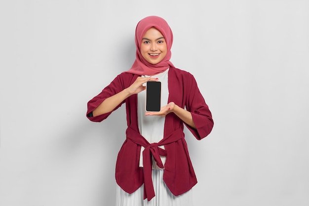 Belle femme asiatique souriante en chemise décontractée et hijab tenant un téléphone portable avec un écran blanc isolé sur fond blanc. Concept de mode de vie religieux de personnes