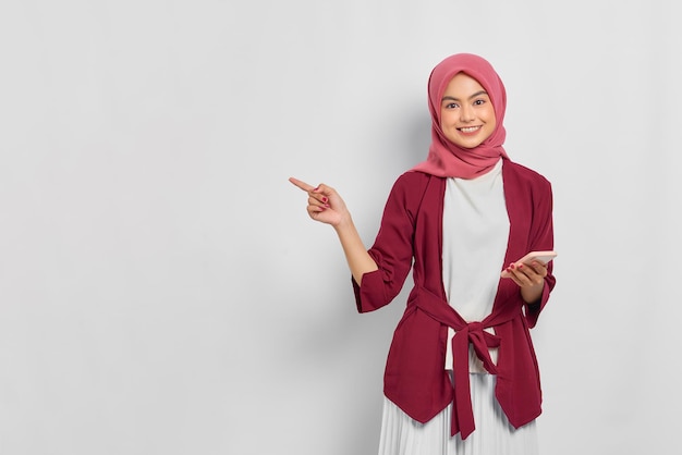 Belle femme asiatique souriante en chemise décontractée et hijab tenant un téléphone portable et pointant du doigt un espace vide isolé sur fond blanc. Concept de mode de vie religieux de personnes