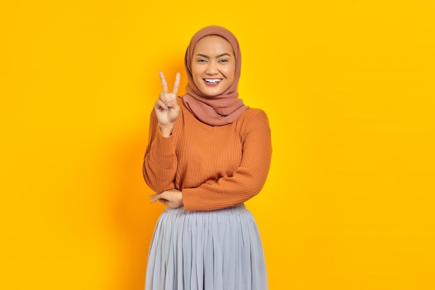 Belle femme asiatique souriante en pull marron et hijab regardant la caméra et montrant le signe de la paix avec le doigt isolé sur fond jaune. concept de style de vie religieux des gens