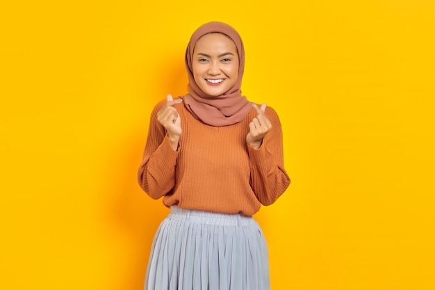 Belle femme asiatique souriante en pull marron montrant le coeur coréen avec deux doigts croisés, exprime la joie et la positivité isolée sur fond jaune