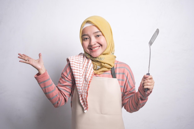 Belle femme asiatique souriante en vêtements roses recouverts de tablier et jilbab tenant une spatule et une écumoire à mailles isolées sur fond blanc