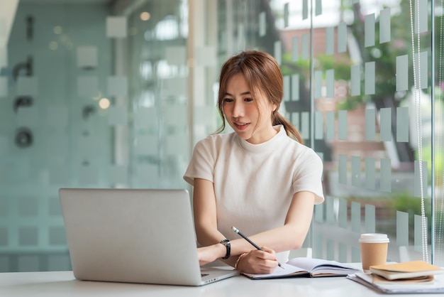 Belle femme asiatique travaillant avec un ordinateur portable et prenant des notes au bureau.