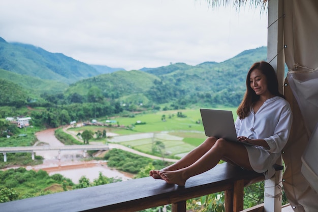 Une belle femme asiatique travaillant et tapant sur un ordinateur portable tout en étant assise sur un balcon avec des montagnes et un fond de nature verdoyante