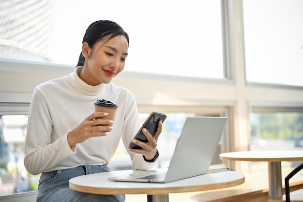 Belle femme asiatique utilisant son smartphone en sirotant un café à distance travaillant dans un espace de coworking