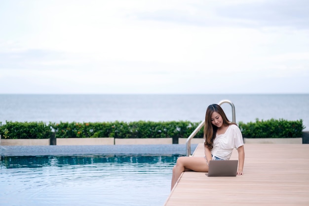 Une belle femme asiatique utilisant et tapant sur un ordinateur portable assis au bord de la piscine