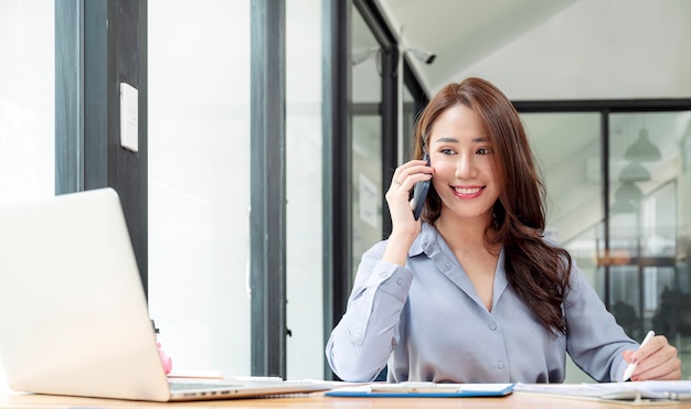 Belle femme asiatique utilisant un téléphone portable tout en travaillant avec un ordinateur portable et de la paperasse au bureau.