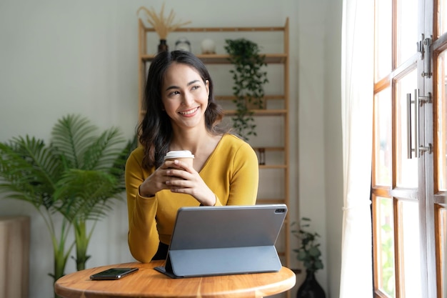 Belle femme asiatique utilise un ordinateur portable en buvant du café et en souriant assis près de la fenêtre à la maison