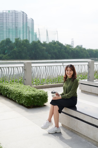 Belle femme assise sur un banc tenant une tasse de café à emporter en plein air.