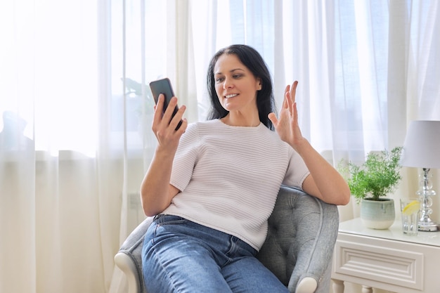 Belle femme assise sur une chaise avec un smartphone parlant en ligne à l'aide d'un appel vidéo