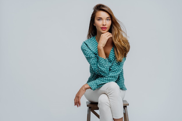 Belle femme assise sur une chaise, tient le menton, porte un chemisier à pois et un pantalon blanc