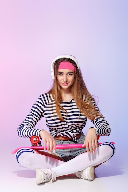 Belle femme assise avec une planche à roulettes et écoutant de la musique sur fond rose