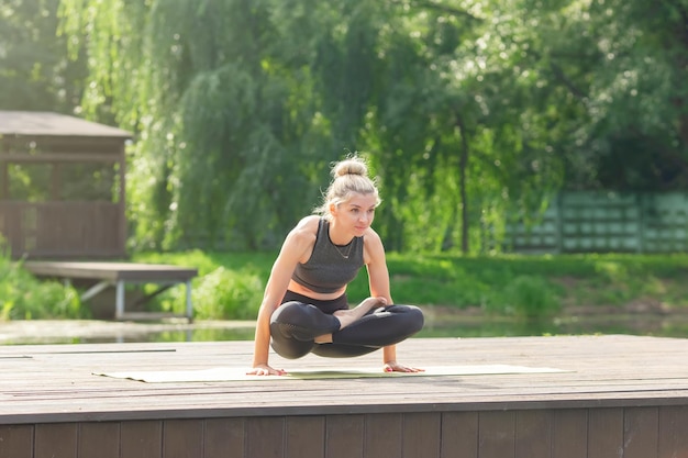 Une belle femme assise sur une plate-forme en bois près d'un étang sur un tapis en été fait du yoga