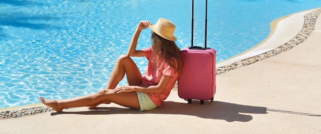 Belle femme assise près d'une valise rose près de la piscine de l'hôtel. Voyage, vacances d'été