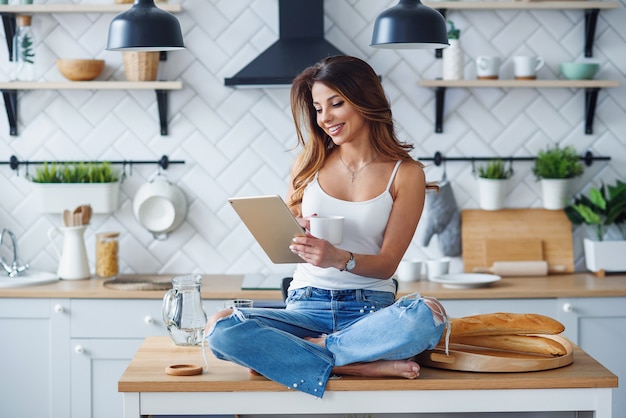 Belle femme assise sur la table et utilise la tablette tactile dans la cuisine confortable le matin à la maison.