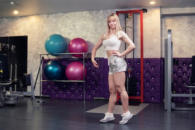 Belle femme athlétique posant dans la salle de gym, le bodybuilder blond sourit et se tient dans un rack pour montrer ses muscles gonflés