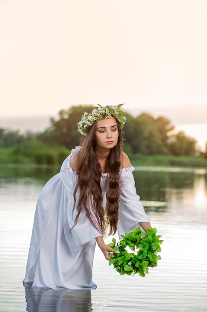 Belle femme au bord de la rivière. Paysanne aux gros seins dans l'eau. Paysage d'été. Femme rustique avec une couronne dans une robe transparente