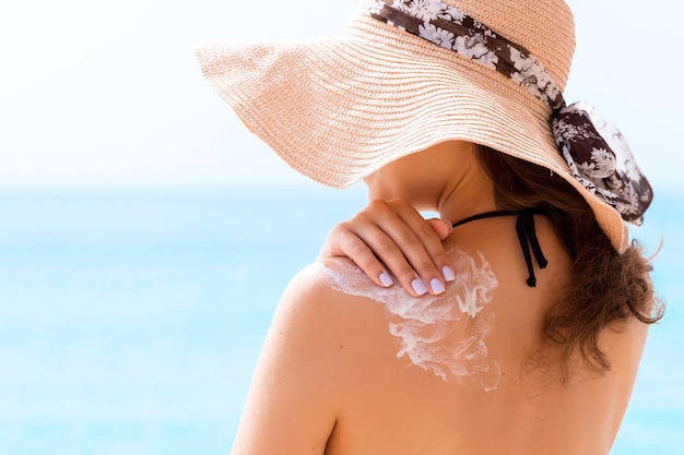 Belle femme au chapeau met de la crème solaire sur son épaule à la plage