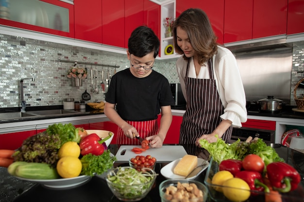 Belle Femme Au Foyer De Femme Asiatique D'âge Moyen Des Années 40 Portant Un Tablier Debout Nouvelle Cuisine De Ton Rouge Et Enseignant Au Fils De 10 Ans à Préparer Des Ingrédients Pour Mélanger Divers Légumes Salade.