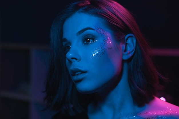 Belle femme au maquillage lumineux avec des paillettes sur son visage dans une pièce avec des lumières bleues et violettes