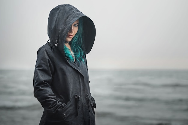 Belle femme aux cheveux bleus se tient près de la mer dans un imperméable avec une capuche et regarde la caméra