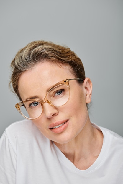 une belle femme aux cheveux blonds et aux lunettes souriant à la caméra sur un fond gris