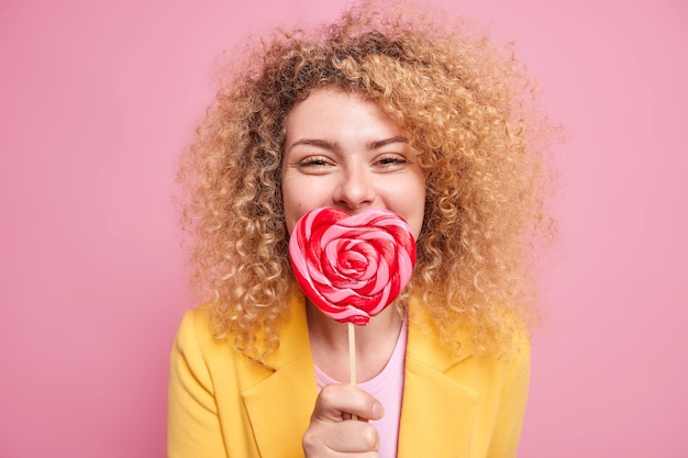 Une belle femme aux cheveux bouclés et touffus garde une sucette en forme de coeur sur la bouche a la dent sucrée aime les bonbons vêtus d'une tenue élégante isolée sur un mur rose. Concept de malbouffe