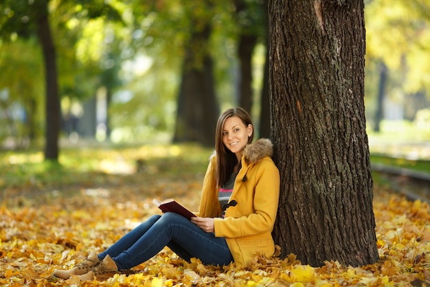 La belle femme aux cheveux bruns souriante et heureuse en manteau jaune et jeans assis sous l'érable avec un livre rouge dans le parc de la ville d'automne par une chaude journée. Feuilles d'or d'automne. Notion de lecture