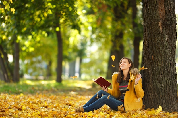 La belle femme aux cheveux bruns souriante et heureuse en manteau jaune et jeans assis sous l'érable avec un livre rouge dans le parc de la ville d'automne par une chaude journée. Feuilles d'or d'automne. Notion de lecture