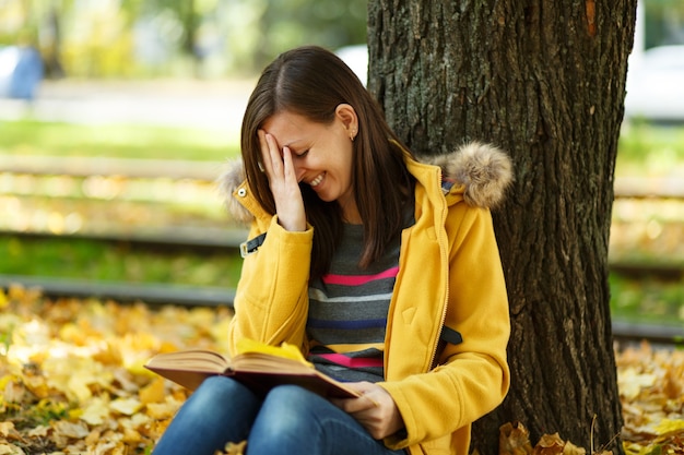 La belle femme aux cheveux bruns souriante et heureuse en manteau jaune et jeans assis sous l'érable avec un livre rouge dans le parc de la ville d'automne par une chaude journée. Feuilles d'or d'automne. Notion de lecture