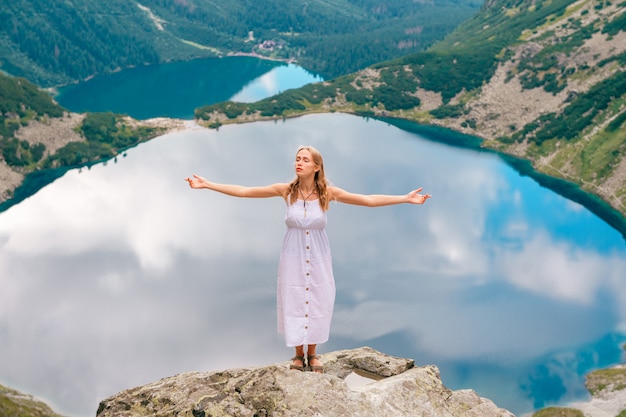 Belle femme aux cheveux longs en robe blanche debout avec les mains écartées dans la pierre dans les montagnes avec des lacs derrière elle