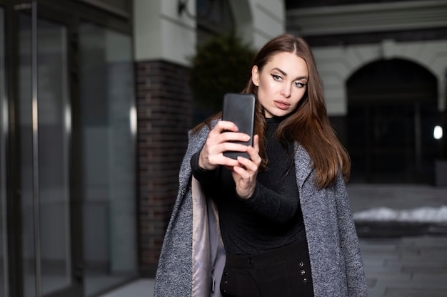 Belle femme aux cheveux longs tient un téléphone dans ses mains et prend un selfie. Une femme d'apparence mannequin est vêtue d'un manteau gris et d'un pull noir. La femme parle au téléphone.