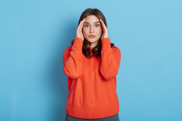 Belle femme aux cheveux noirs, garde les mains près des yeux, porte un pull orange décontracté, posant isolé sur un mur bleu, regarde avec de grands yeux à la caméra, en gardant le geste de se cacher.