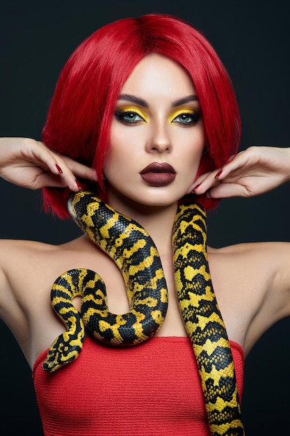 Belle femme aux cheveux rouges et un serpent python jaune autour du cou. Maquillage professionnel des yeux de beauté lumineuse jaune, rouge à lèvres rouge bordeaux