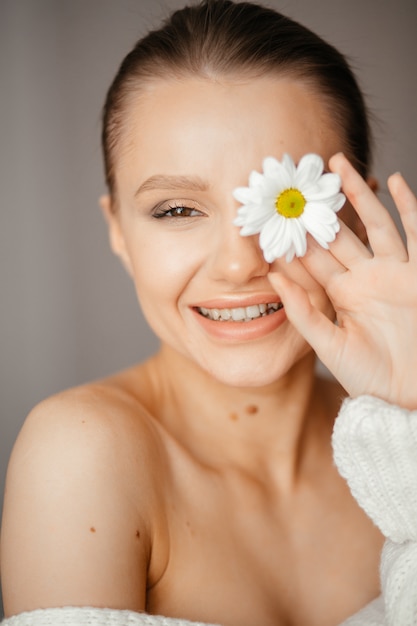 Belle femme aux yeux fermés dans un pull blanc rit et apporte une fleur blanche à ses yeux